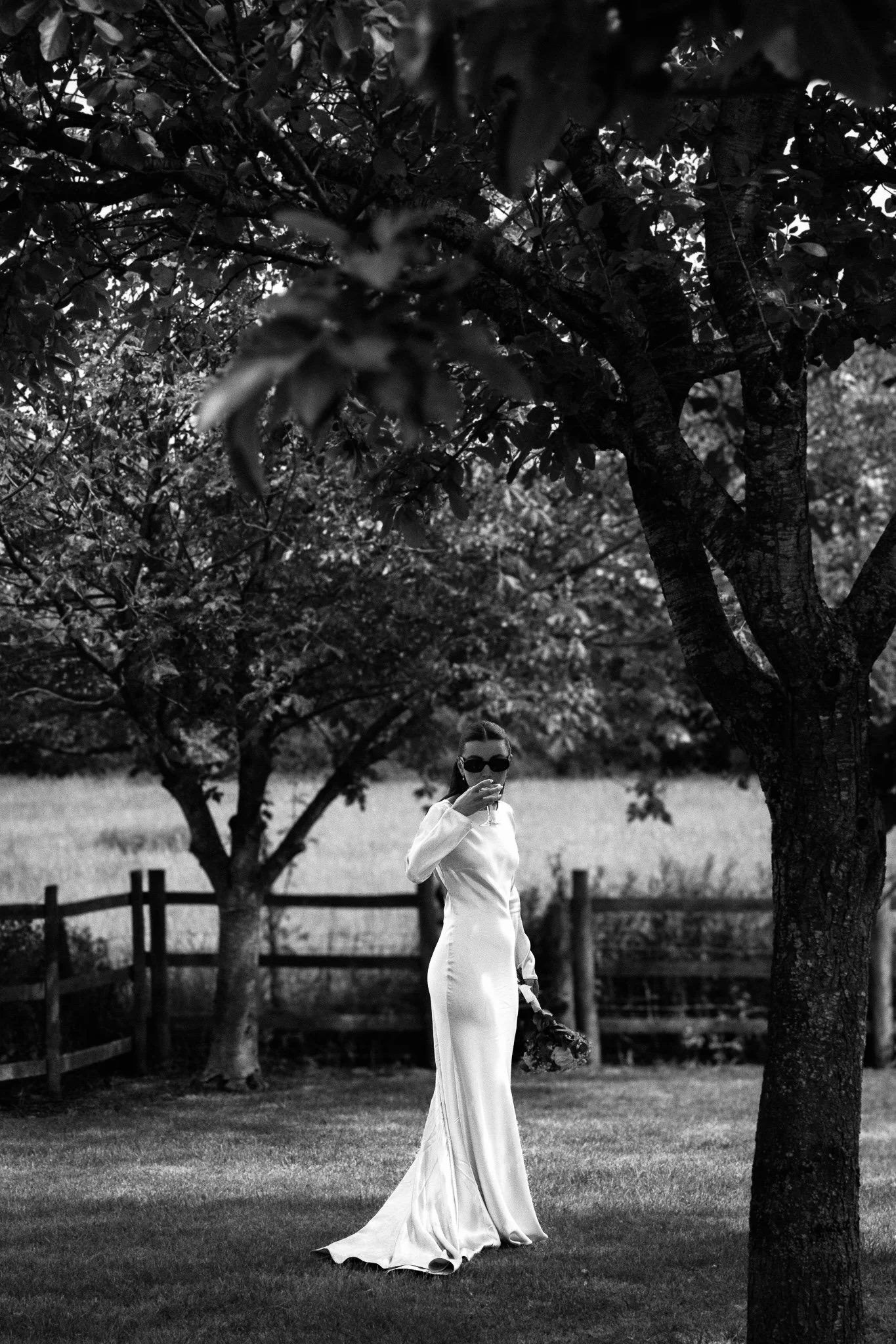 A bride in a long satin dress standing beneath trees in a meadow, glass in hand